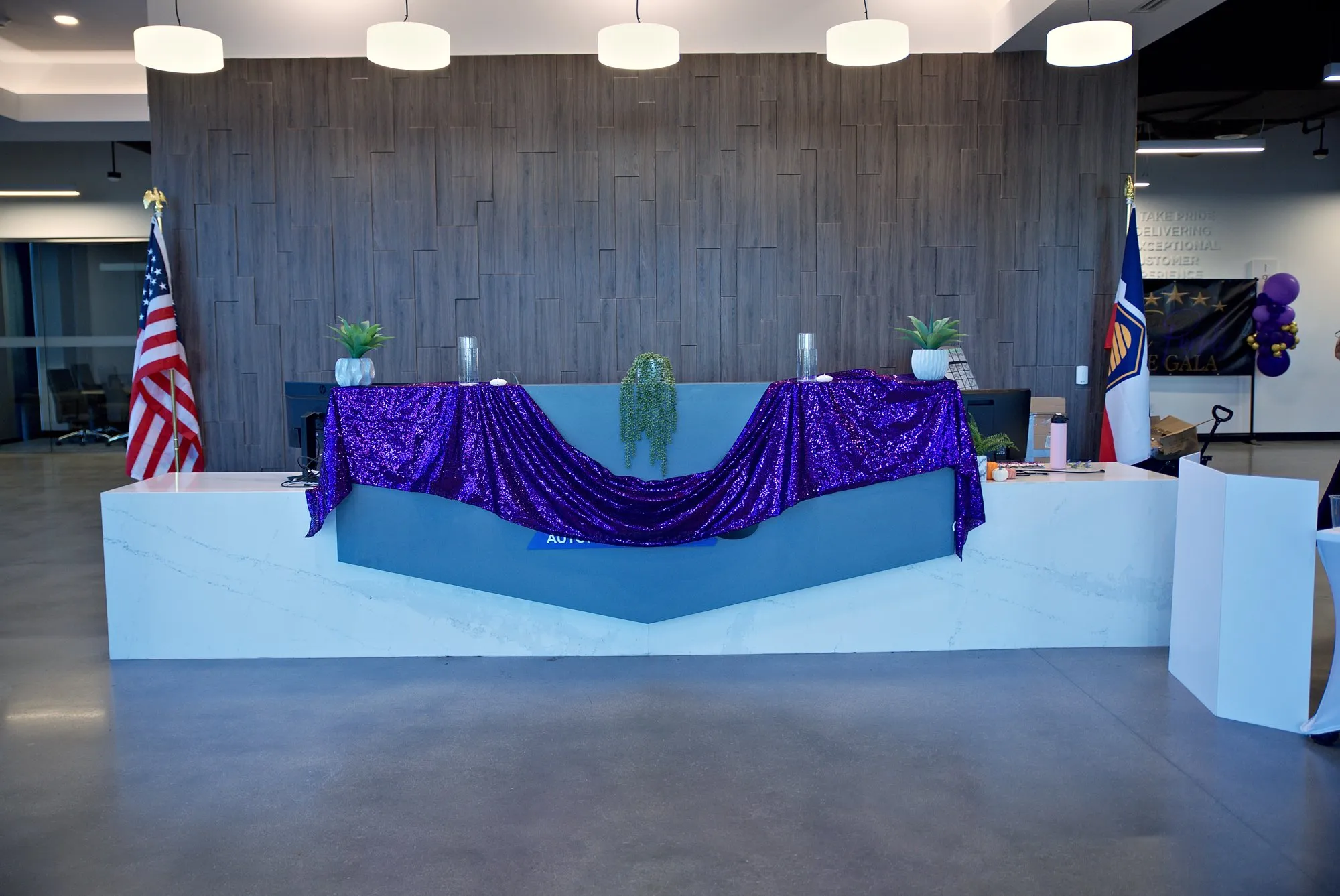 The reception desk head-on, purple sequin drape between two flags under pendant lights