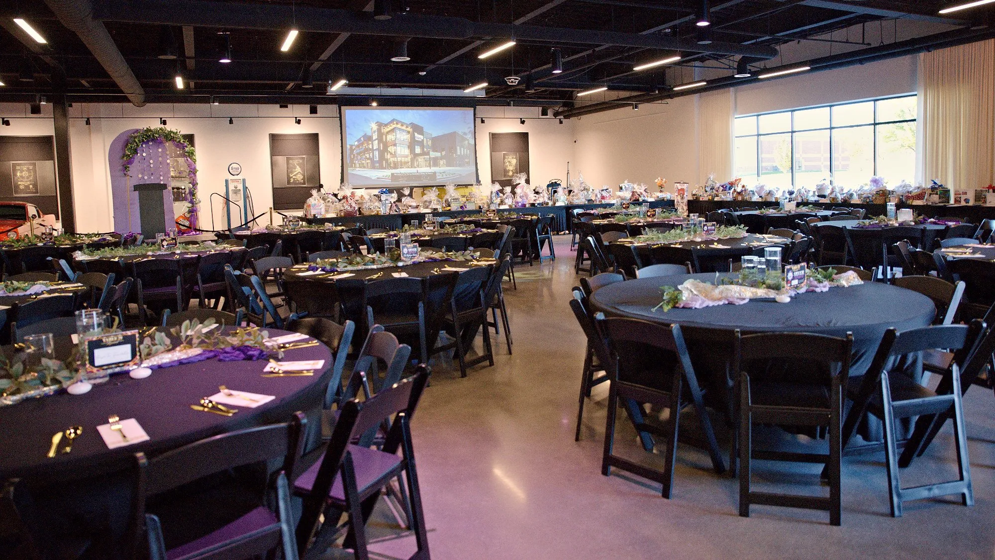 The gala room set before doors open: dressed round tables under ceiling lights, a projected screen, and tall windows